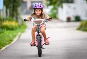 Children learning to drive a bicycle on a driveway outside.